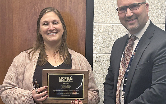Jessica is holding here award in hand as she stands beside here school superintendent. Background is an off white colored cinderblock wall and a wooden door.