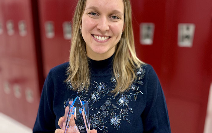 Jennifer Kane stands in front of red school lockers holding her award in her hands and smiling at the camera.